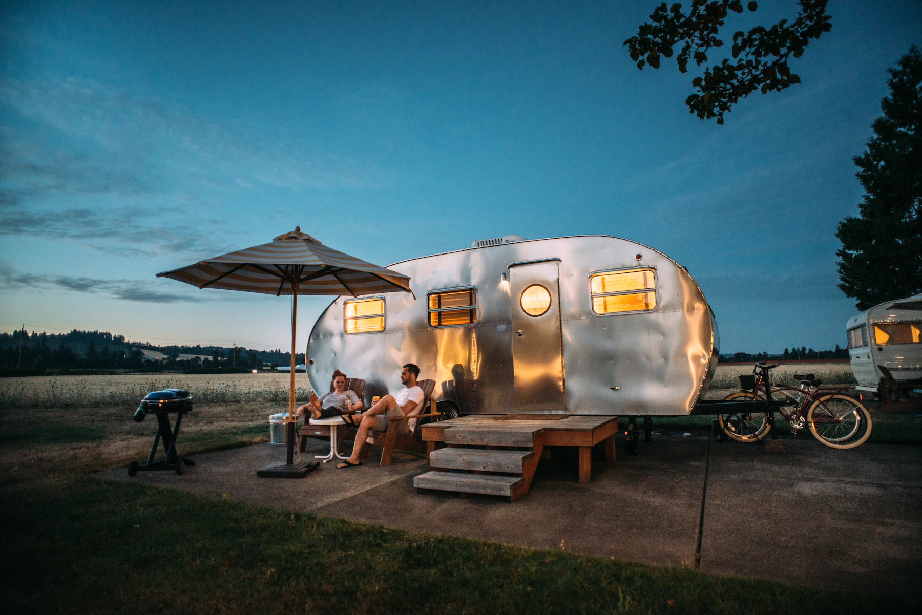 A vintage caravan lit from inside at dusk, with two people sitting outside under a parasol